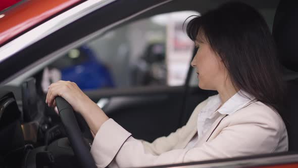 Side View of Confident Brunette Woman Sitting on Driver's Seat Choosing Vehicle in Car Dealership