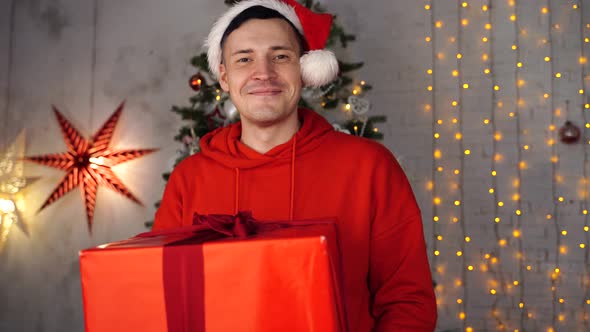 Young Handsome Man in Santa Claus Hat Dancing with Big Gift on Background of Christmas Tree. Happy