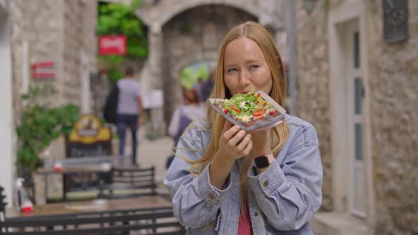 A Young Woman Eats a Delicious Pizza on Black Bread Standing on a Street of an Old European Town