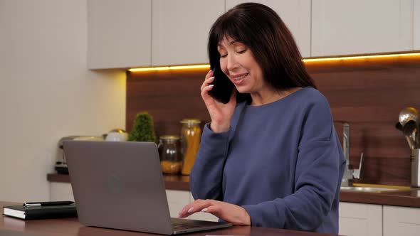 Smiling Businesswoman Talking on Mobile Phone Works for Laptop in Home Kitchen