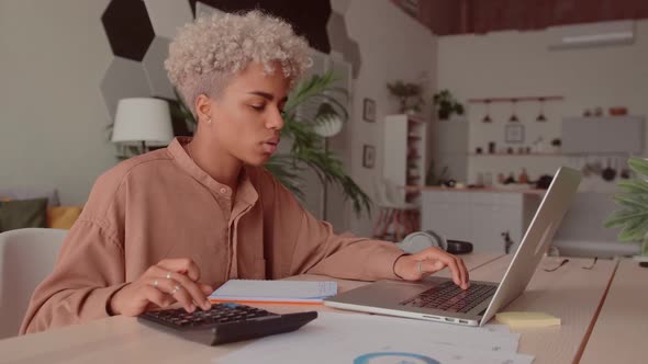 African Woman Calculates Costs Using Calculator and Laptop Sit at Home Desk