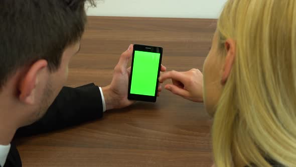 Two Office Workers, Man and Woman, Sit at A Desk and Scroll at A Smartphone with A Green Screen