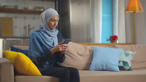 Portrait of Arabic Woman in Hijab Using Smartphone Sitting on Sofa Relaxing at Home.