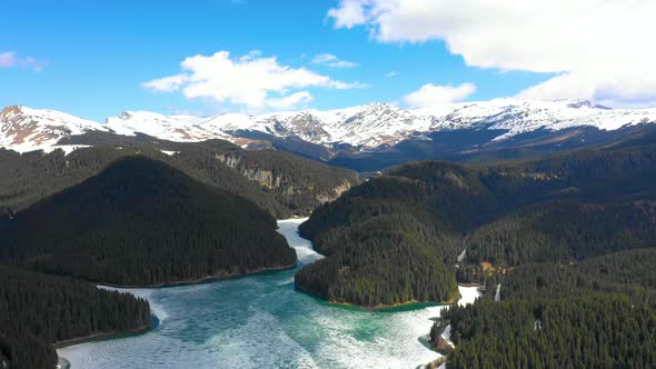 Aerial view of a frozen mountain lake surrounded by forest and snowy mountains in spring season.