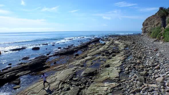 Side view tracking shot of a young man running on a rocky ocean beach shoreline.