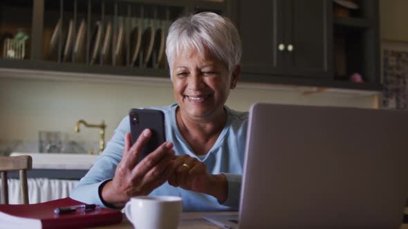 Happy senior mixed race woman using smartphone and laptop in kitchen