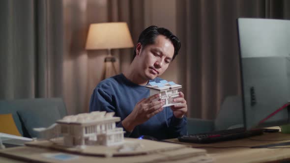 Asian Male Engineer Holding And Looking At House Model With Solar Panel While Working On Desktop