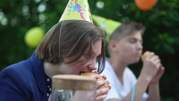 Side View Portrait of Teenage Caucasian Girl in Party Hat Biting Chewing Sandwich Looking at Camera