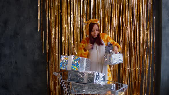 Woman in Pajamas Puts Gifts in Shopping Cart Near Wall with Golden Tinsel