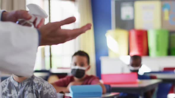 Diverse teacher disinfecting hands in classroom, with schoolchildren sitting wearing face masks