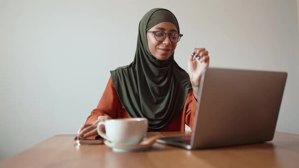 Cheerful Muslim woman wearing eyeglasses working on laptop