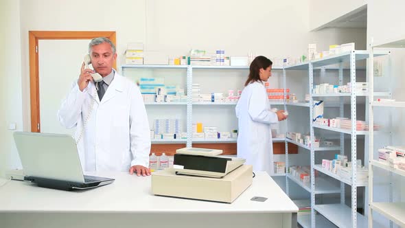 Pharmacist talking on the phone in a pharmacy while using a laptop