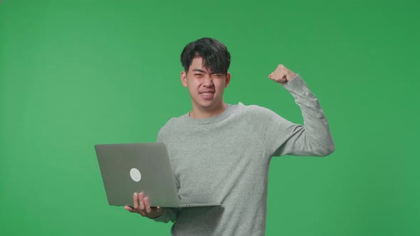 A Smiling Asian Man Flex Muscle While Typing On Computer In The Green Screen Studio