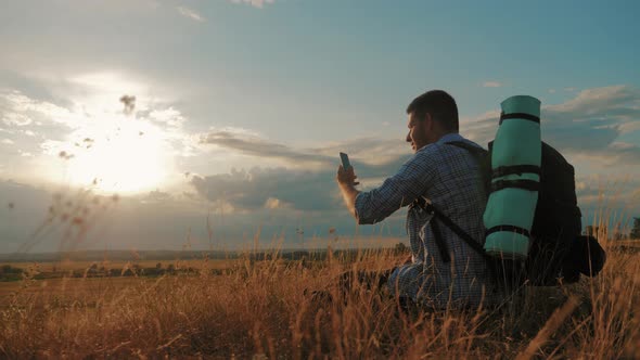 A Young Tourist Man with a Backpack Uses a Mobile Phone. In the Rays of the Setting Sun. Travel
