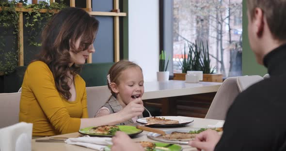 Happy Young Mother Feeding Cute Charming Daughter with Pizza in Restaurant