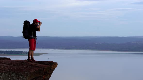 man traveler with backpack using camera taking a photo on the edge of cliff