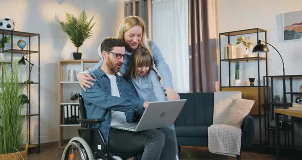 Man in Wheelchair Applying Laptop Together with His Daughter and Pretty Joyful Wife at Home