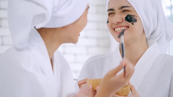 Beautiful young woman in white bathrobe applying a revitalizing black mask onto her friend's face.