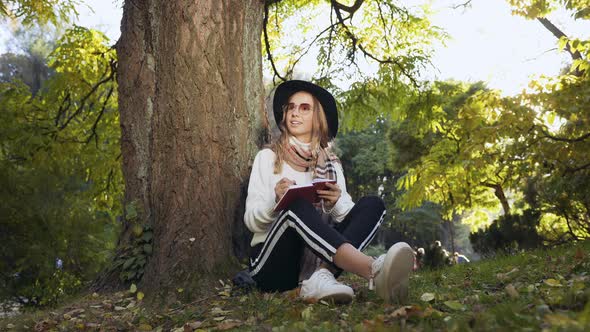 Young Woman in the Hat and Sunglasses Sitting Near Tree in Green Park