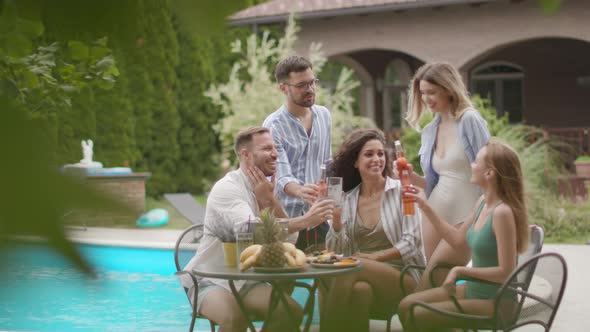 Group of happy young people cheering with cider by the pool in the garden