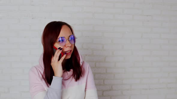 Young Woman in Glasses Speaks on Smartphone Standing Near White Brick Wall