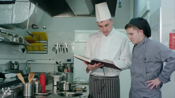 Chief Chef Holding Recipe Book and Explaining Something To His Young Trainee