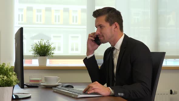 An office worker in a suit in front of a computer talks on a smartphone