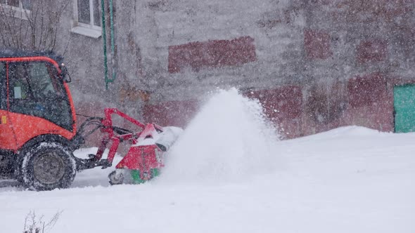 Defocused Red Tractor with Snowplow Cleaning Snow Sidewalk at Winter Daylight Blizzard