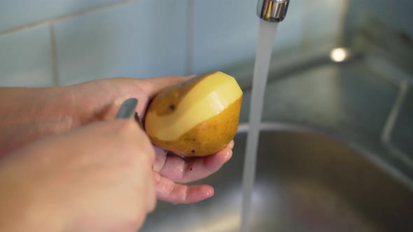 Woman Peels Ripe New Potatoes with Vegetable Peeler Over Silver Sink Near the Flowing Water in the