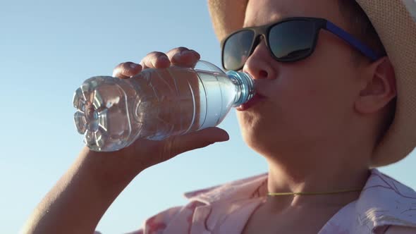 Boy in a Hat and Sunglasses Drinks Water From a Bottle on the Beach