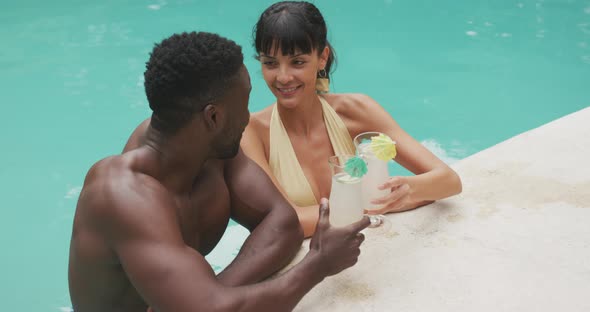 Happy diverse couple wearing swimming suits and drinking drinks at swimming pool in garden