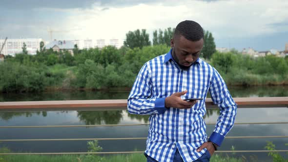 Black Man Browses Phone Looks at Camera and Smiles Stands on Waterfront in Park.