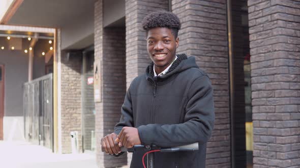 A Young African American Man in Casual Dark Clothes with a Scooter Stands Near a Building with a