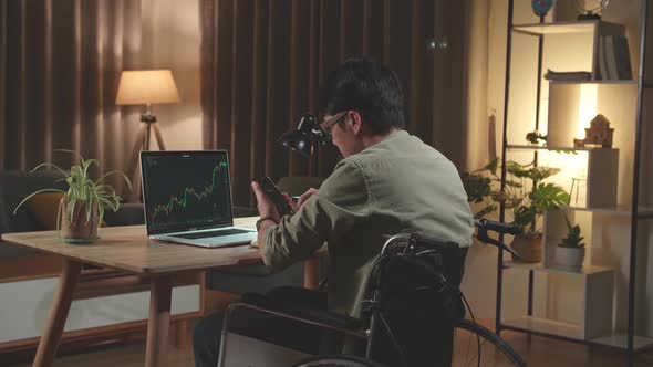 Asian Man Trader Investor Sitting In A Wheelchair Using Phone And Laptop Trading Cryptocurrency