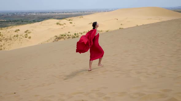 Sexy Brunette Woman in a Red Satin Long Dress Walks on Sand Dunes in the Desert