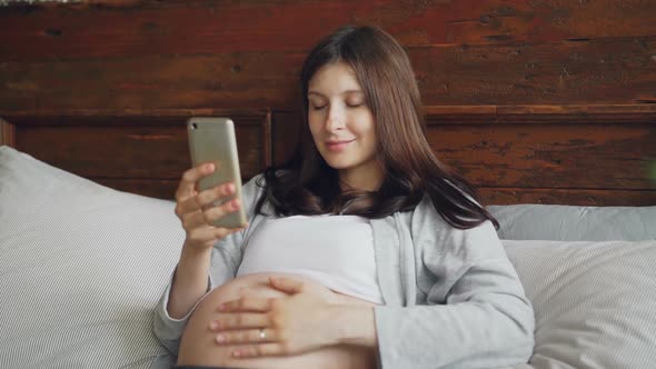 Smiling Pregnant Young Lady Is Using Smartphone and Caressing Her Tummy Resting in Bed in Modern