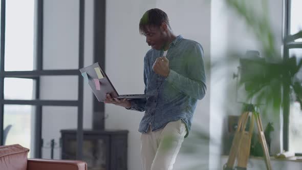 Satisfied African American Young Man Dancing in Slow Motion in Home Office Holding Laptop in Hand