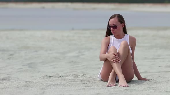 Languid Woman Sitting Cross-legged on Sand Touching Skin