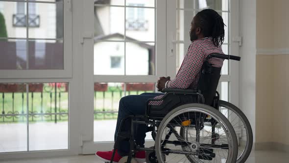 Wide Shot Thoughtful Disabled African American Man in Wheelchair Sitting Indoors Looking Out the