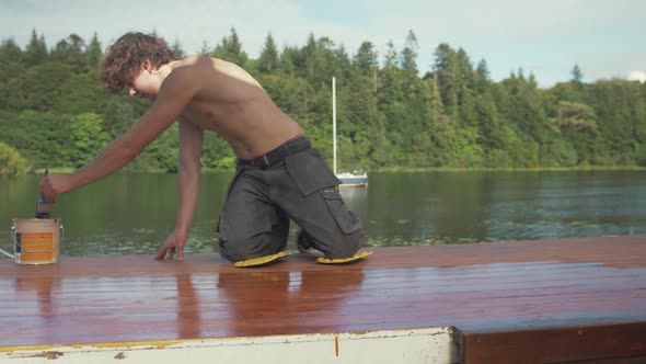 Young man painting wooden boat cabin roof planking with protective exterior paint.