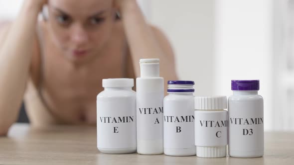 Row of Vitamin Bottles on Table with Blurred Caucasian Young Woman at Background Thinking