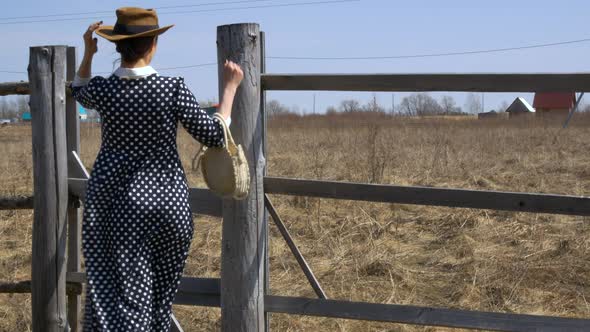 Pretty Girl in a Hat and White Straw Handbag Standing Near the Fence on a Windy Day