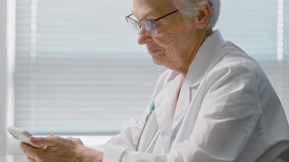 Cheerful senior lady doctor with spectacles texts message