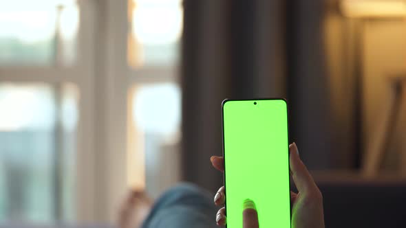 Woman at Home Lying on a Sofa and Using Smartphone with Green Mock-up Screen in Vertical Mode. Girl