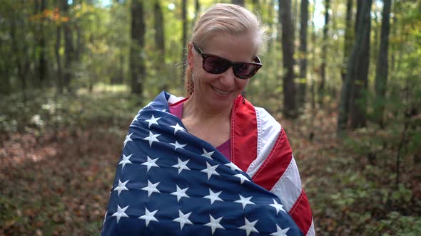 Closeup of pretty, blonde woman wrapping an American flag around her and smiling in a sunlit forest.