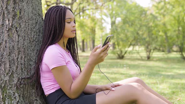 A Young Black Woman Listens To Music on a Smartphone with a Smile As She Sits Under a Tree in a Park