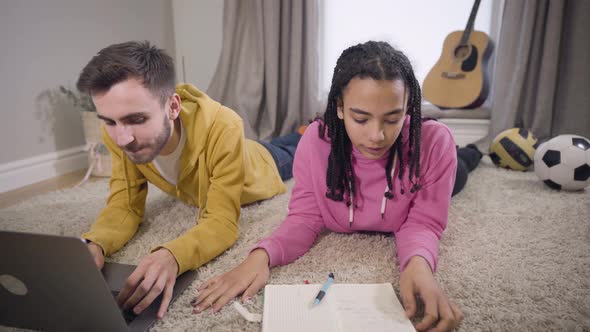 Concentrated African American Girl Writing and Putting Hand on Floor As Caucasian Boy Touching Her