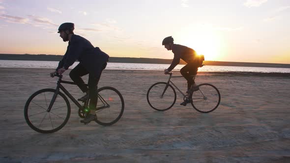 Two Young Men Riding Bicycles on the Beach on the Background of an Orange Sunsetting Sky