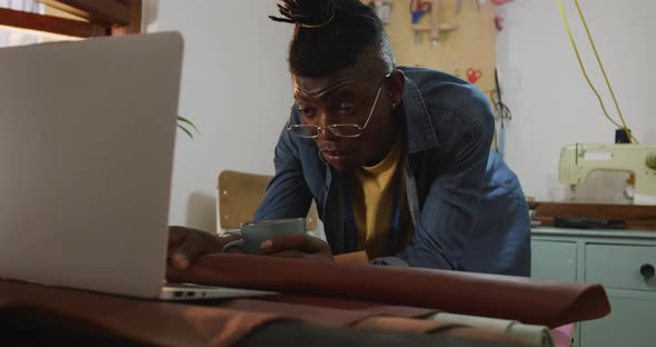 African american craftsman wearing glasses drinking coffee and using laptop in leather workshop