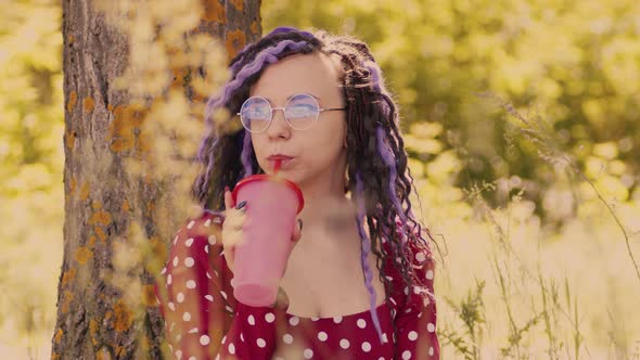 Young Woman in Glasses Red Dress Drinking Refreshing Beverage Sitting Near Tree in Sunny Windy